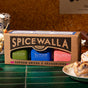A boxed Spicewalla Hot Chocolate Drink Mix Collection sits on a green tile counter near a cinnamon roll and popcorn, featuring three tins: Azteca, Classic, and Cinnamon Chili Hot Chocolate.
