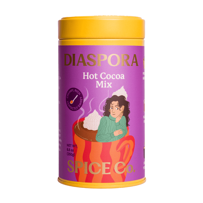 A purple and yellow tin labeled Hot Cocoa Drink Mix from Diaspora Spice Co. features an illustration of a woman in a green sweater sitting in a red mug topped with whipped cream, capturing the warmth of a hot cocoa blend crafted with single-origin spices and sweetened with Madhur Jaggery.