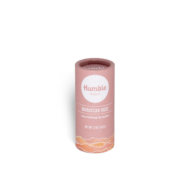 A pink cylindrical tube of Humble Brands Lip Balm - Paperboard stands upright on a white background, featuring a white label with a wave design and details including the use of organic coconut oil for moisturizing care.