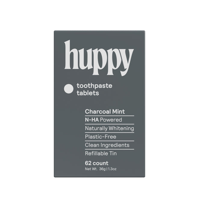 A dark gray Huppy Toothpaste Tablets with Tin in Charcoal Mint, featuring N-HA for cavity protection, natural whitening, clean ingredients, refillable tin, plastic-free packaging, and 62 tablets for a sustainable brushing routine.