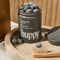 A tin labeled Huppy holds sustainable Toothpaste Tablets, some scattered nearby. A wooden toothbrush with beige bristles rests on a round beige tray, while the tin lid sits to the side.