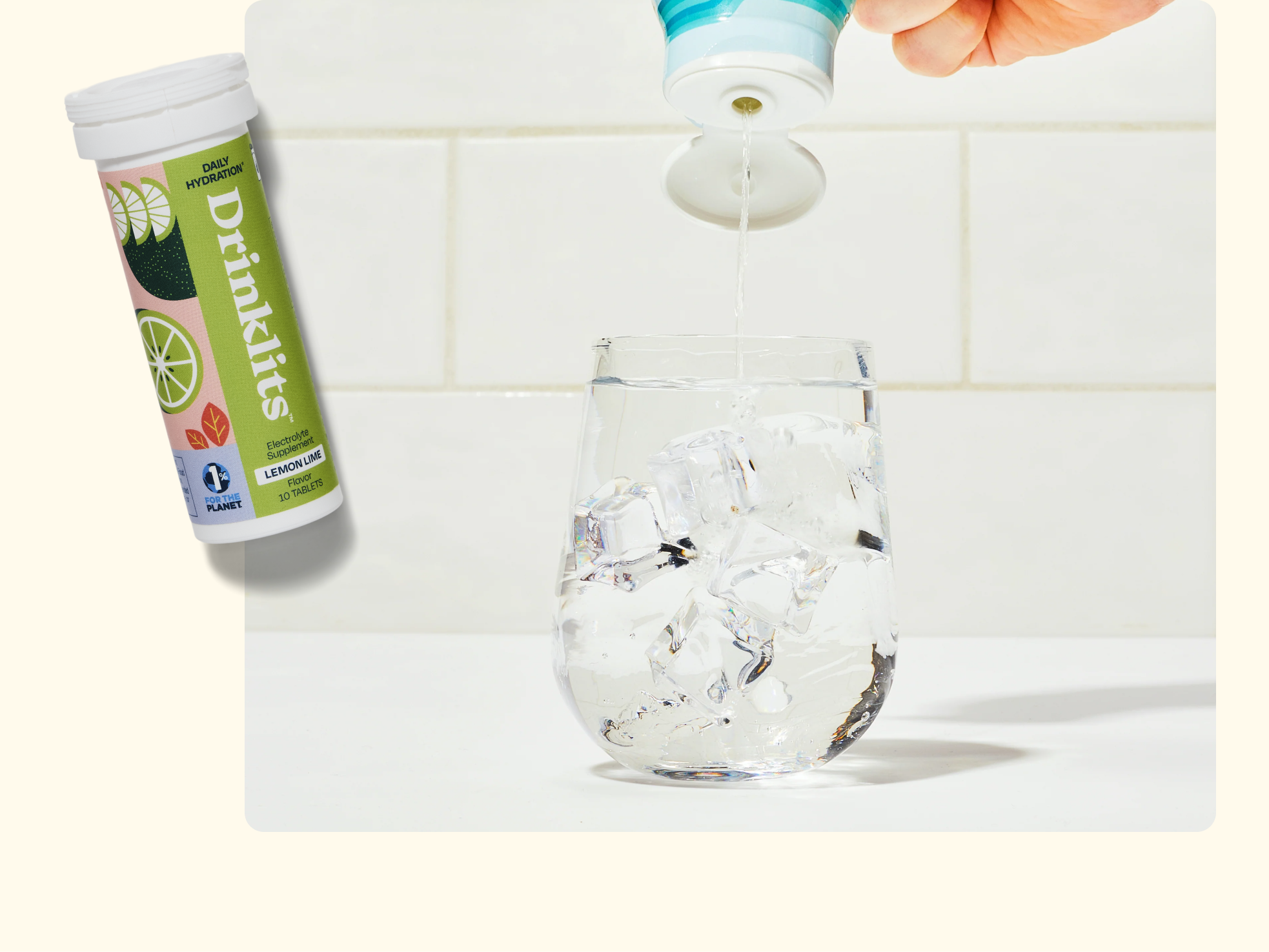 A hand pours liquid from a blue bottle into a glass filled with ice. Next to the glass, there is a Drinkits tube showing lemon and lime flavor images. The background is a white tiled wall.