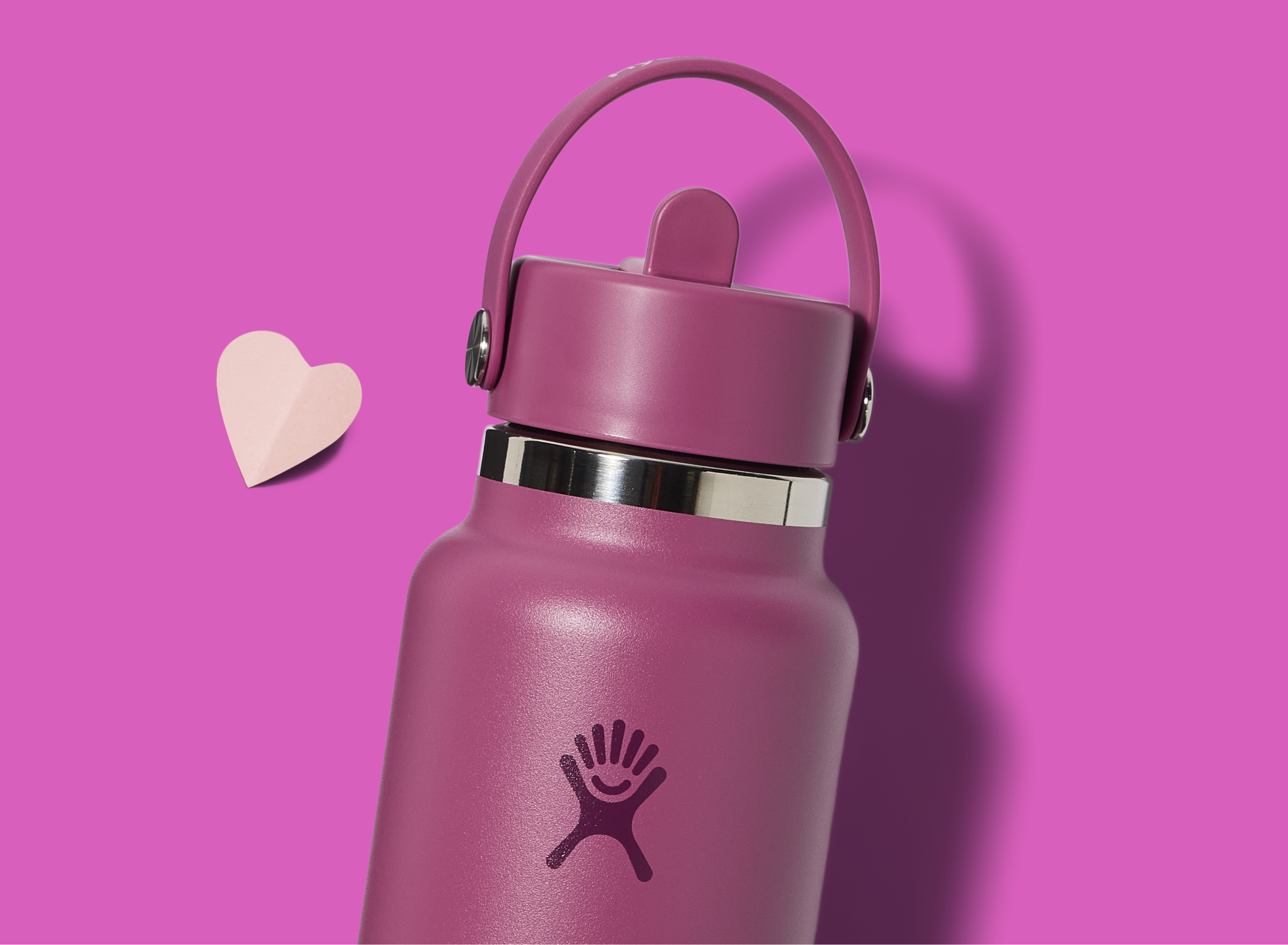 A pink insulated water bottle with a handle and a white smiling logo on the front sits on a bright pink background next to a small paper heart.