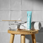 A tube of Davids Hydroxi™ Premium toothpaste - Whitening + Enamel Health w/nano-hydroxyapatite, a toothbrush with paste, a glass of water, and a rolled white towel are neatly arranged on a wooden stool against a tiled wall, emphasizing natural enamel health.
