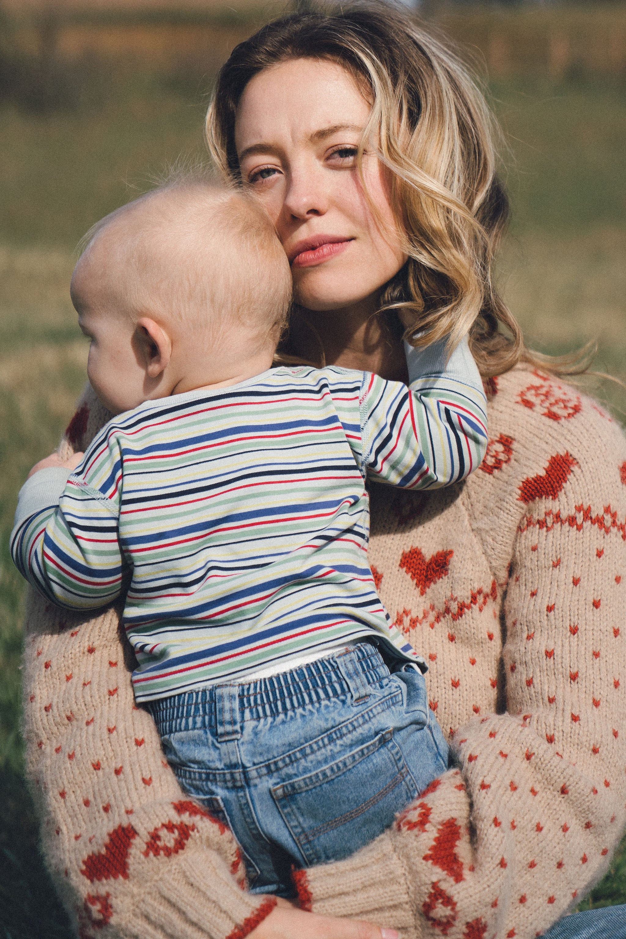 A woman with wavy blonde hair and a beige sweater with red hearts holds a baby in her arms outdoors. The baby, in a striped shirt and jeans, faces away while the woman looks at the camera.