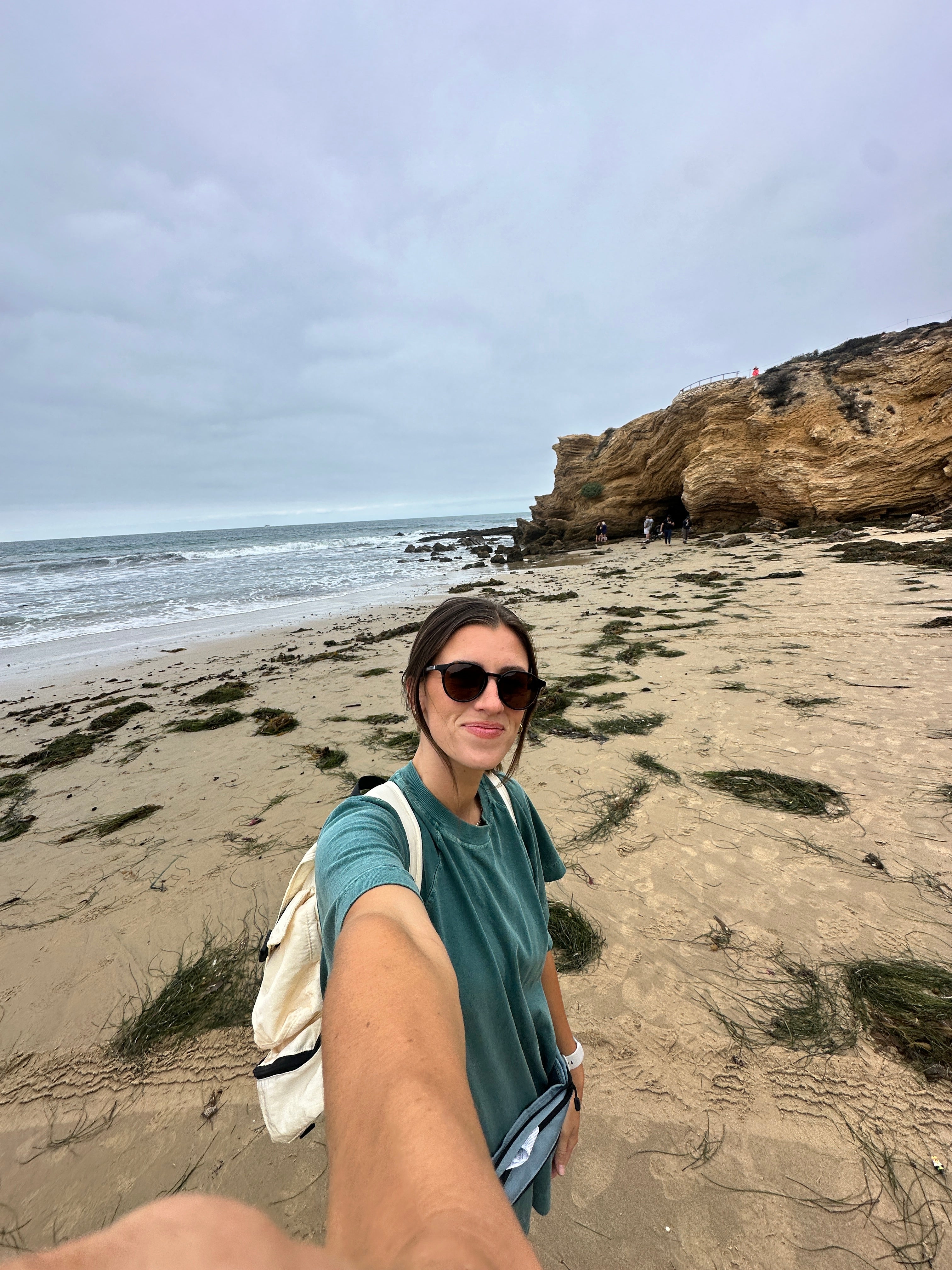 A person wearing sunglasses and a green shirt takes a selfie on a sandy beach with rocky cliffs and seaweed. The ocean and cloudy sky are in the background.