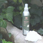 A silver spray bottle of grab greens Room and Fabric Freshener - Gardenia, enhanced with essential oils and phthalate-free, sits on a tree branch amid green leaves. A white flower is next to it, offering a natural odor-neutralizing experience.