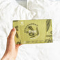 A hand holds a green box of Stoneworks Dryer Sheets - Olive Leaf by grab green, featuring olive branch illustrations. Made from recycled paper, it combats static cling with 50 sheets against a blurred white bedspread background.