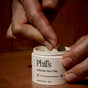Hands scoop Phil's Icelandic Hair Clay from a white jar, highlighting its compostable packaging and natural formula with Icelandic kelp and clay for effortless hair styling.