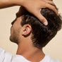 A person with short dark hair styled naturally using Phil's Icelandic Hair Clay stands out in a white shirt against a plain beige background, resting their hand on the back of their head.