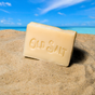 A beige Bar Soap from Old Salt rests on sandy beach, with the blue ocean and sky stretching behind it.