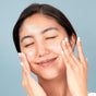 A woman with clear skin and closed eyes smiles softly, gently touching her cheeks against a light blue background, highlighting the soothing effects of FaceTory's Calming Face Sheet Mask—perfect for sensitive skin.