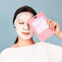 A woman with her eyes closed smiles while holding up a FaceTory Calming Face Sheet Mask packet for sensitive skin against a light blue background.