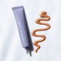 A lavender tube labeled Bronzing Drops from Indie Lee rests beside a wavy swipe of shimmery bronze liquid, perfect for a summer glow, on a white surface.