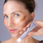 A blue-eyed, light-skinned woman smiles while holding a tube of Indie Lee Bronzing Drops near her face. She has a swipe of the bronzing serum on her cheek, giving her skin a radiant summer glow.
