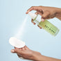 A hand holds a cotton pad while another sprays mist from a green Indie Lee Squalane Facial Oil - Travel Size bottle, made with olive-derived squalane and known for clean skincare, onto it against a light blue background.