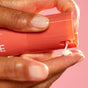 A close-up of a hand pressing Indie Lee’s Retinol Alternative Cream pump with coral-colored packaging, dispensing the anti-aging cream onto another hand against a pink background.