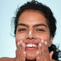 A smiling woman with curly hair applies Indie Lee Purifying Face Wash - Travel, a natural cleanser and makeup remover, to her face with both hands against a light blue background.