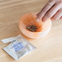 A hand reaches into a peach-colored bowl of brown tablets, with an open Indulgence Relief Sachet - Sample by WTHN nearby—Chinese herbs formulated to support liver detox after alcohol metabolism.