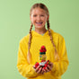 A smiling girl in a yellow hoodie with braided hair holds a bottle of MegaFood Iron Energy Gummies, surrounded by fresh strawberries, against a light green background—delicious support for energy.