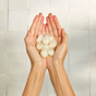 A pair of hands holds translucent, round white Dew Bomb Glowy Hydra-Milk Starter Kit capsules from IZZY Zero Waste Beauty against a light tiled background.