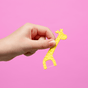 A hand holds a Jack N Jill Fairy Floss Floss Pick—yellow, giraffe-shaped, and ideal for child dental care—against a solid pink background.