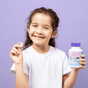 A smiling young girl in a white shirt holds a Tooth Sparkles Chewable Teeth Cleaning Tablet in one hand and a Jack N Jill bottle in the other, standing against a light purple background—ideal for highlighting kids’ oral care supplements.