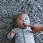 A baby in a sheep-patterned onesie lies on a gray furry carpet while being fed with the Jack N Jill Silicone Finger Brush 2-PACK STAGE 1, supporting good baby oral hygiene.