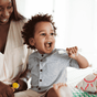 A smiling young child in a plaid shirt holds a Jack N Jill Silicone Toothbrush STAGE 2 and opens his mouth wide, while an adult beside him smiles and holds a yellow toy. They are seated on a patterned bedspread.