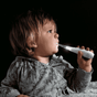 A young child with light brown hair, dressed in a grey animal-print shirt, holds the Jack N Jill Tickle Tooth Sonic Toothbrush with a glowing LED to their mouth against a dark background.