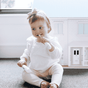 A smiling baby in a headband and white clothes sits on the floor, holding and chewing on a Jack N Jill Tickle Tooth Sonic Toothbrush, with a toy house visible in the background.