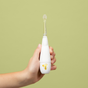 A hand holds a white Jack N Jill Tickle Tooth Sonic Toothbrush for kids, featuring a small giraffe on the handle, against a plain light green background.