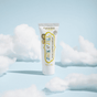 A tube of Jack N Jill Natural Toothpaste stands upright on a blue background, surrounded by fluffy cotton props. The fluoride-free, flavor-free toothpaste features a yellow and white design.