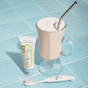 A tube of Jack N Jill Natural Toothpaste and a white toothbrush sit next to a glass of frothy milkshake with a metal straw, all arranged on a blue tiled surface.