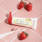 A tube of Jack N Jill Natural Toothpaste rests on pink tiles with three strawberries and a white toothbrush with pink, fluoride-free toothpaste nearby.