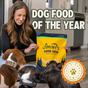 A woman smiles while holding a bag of Jiminy's Good Grub Dry Dog Food (Allergy Relief, Healthy Skin & Coat) by Jiminy's, surrounded by three dogs. Text reads "Dog Food of the Year" with a 2024 Pet Innovation Award badge.