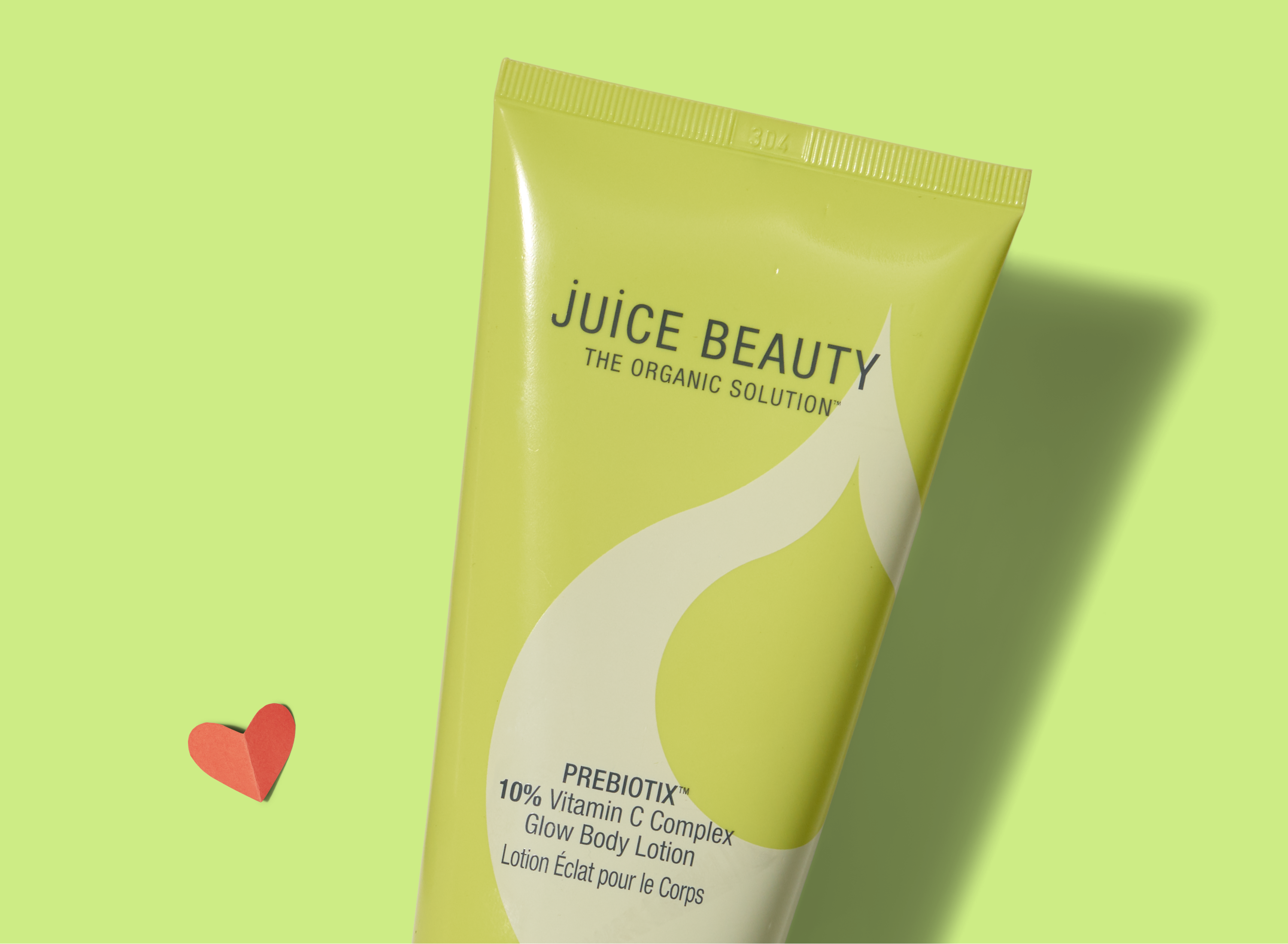 A green tube of Juice Beauty Glow Body Lotion lies on a light green background with a small red paper heart nearby. The label highlights prebiotics and 10% vitamin C complex.
