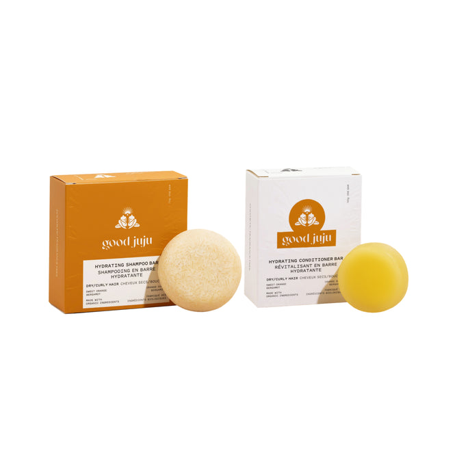 The Hydrating Haircare Set by Good Juju Body & Home includes a beige shampoo bar in an orange box and a yellow conditioner bar in a white box, all labeled “Hydrating” for dry, curly hair. Products are shown on a white background.