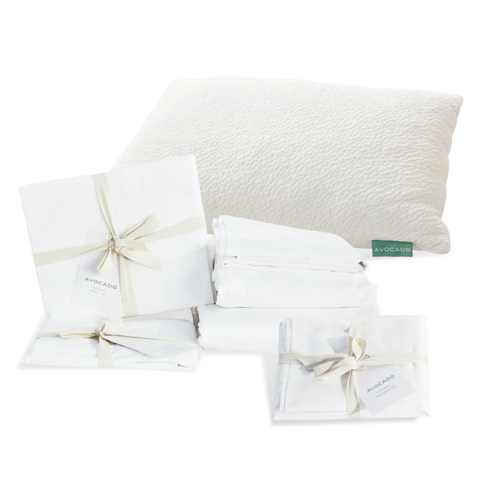 A fluffy white pillow with a green "Avocado" tag and neatly folded white sheets, bundled with ribbons and tags, are shown on a white background—Grove Collaborative’s Highest Impact Bedroom Swap Set: Everyday Sleep Essential.