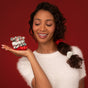 A smiling woman with curly hair in a side ponytail holds the Holiday Ruched Satin Scrunchies Gift Set by Kitsch, wearing a white fuzzy top against a deep red background.