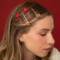 A woman with light brown hair wears three Kitsch Metal Bobby Pins Gift Bow Set hair pins—each topped with small red and gold gift bows—against a solid red background.