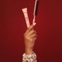 A hand with manicured nails holds the Kitsch Hair Taming Sleigh All Day Gift Set and a styling brush against a red background, wearing a beige scrunchie—perfect for sleek hairstyles.