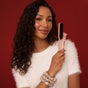 A woman with curly hair in a white fuzzy top smiles and holds the Kitsch Hair Taming Sleigh All Day Gift Set—stylish pink brush and product—in front of a red background, ready to create sleek hairstyles.