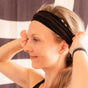 A woman with light brown hair smiles as she adjusts the KOOSHOO Organic Twist Headband on her head, wearing a fitness tracker on her wrist and standing before a black and white patterned background.