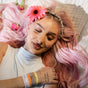 A young woman with long, wavy pastel pink hair lies on a bed, eyes closed, wearing KOOSHOO Organic Hair Ties as colorful bracelets on her wrist and an ethically made, sleeveless white top.