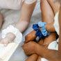 Two people sit side by side on a light blanket with KOOSHOO Scrunchies—one white, one dark blue, and one light blue—on their wrists. One reads a book. Their faces are not shown.