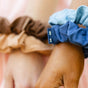 Two hands, each wearing two KOOSHOO Scrunchies in brown, beige, blue, and light blue organic cotton. The background features a softly blurred pastel design.