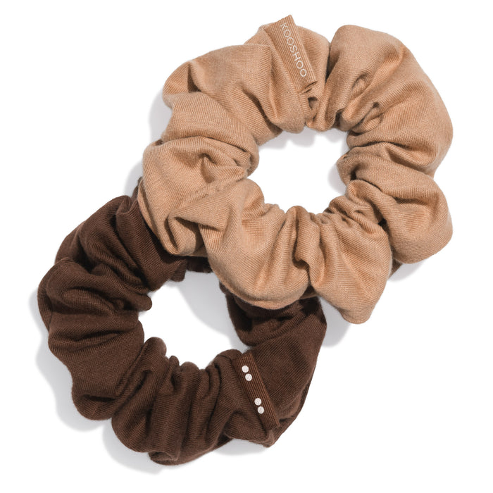 Two KOOSHOO Scrunchies made from organic cotton—one light beige with a sewn label, one dark brown—are intertwined on a white background, each featuring small decorative details.