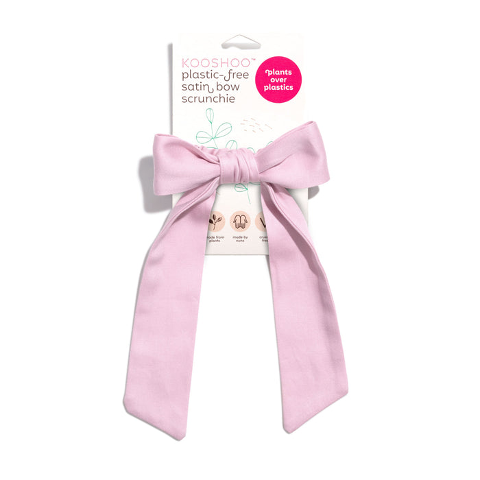 The KOOSHOO Satin Bow Scrunchie, made from plant-based satin and organic cotton, is shown on its packaging with a pink circle that reads “plants over plastics,” emphasizing its zero-plastic design.