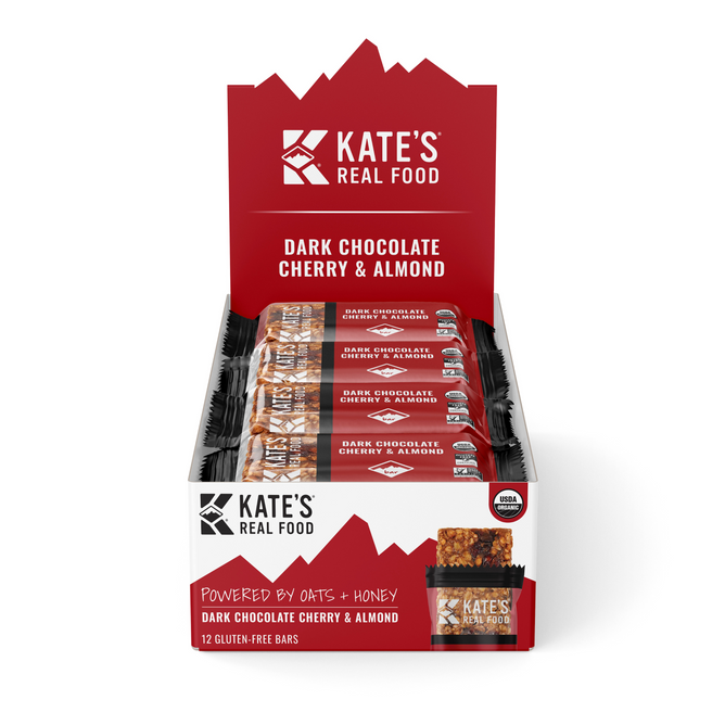 A display box of Kate’s Real Food Organic Energy Bar - Dark Chocolate Cherry & Almond provides sustained energy. Each box contains several individually wrapped bars in red packaging with the brand’s real food imagery.